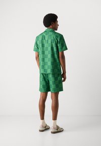 Zöld mintás, rövid ujjú ing és hozzáillő rövidnadrág, puha, sima anyagból, geometrikus mintával. Barna sportcipővel viselve.
