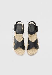 Sandali in pelle nera con cinghie incrociate, suola in juta intrecciata e chiusura con fibbia. Suola interna testurizzata e dettagli di cucitura sottili.
