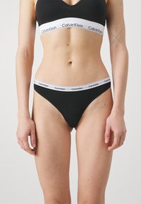 Črne bombažne tangice z belim elastikom na pasu, na katerem je napis "Calvin Klein". Gladka tekstura, minimalističen dizajn, nizko rezanje.