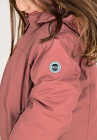 Rosa isolierte Jacke mit einer Reißverschlusstasche und runder Logodetail auf dem Ärmel. Die Jacke bietet eine glatte Textur und ein Kapuzendesign.