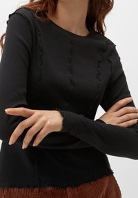 Musta ribattu pitkähihaine paita, jossa on aaltoilevat reunat kaula-aukossa ja hihoissa. Paitaan kuuluu istuva malli ja teksturoitu pinta.