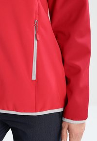 Rød softshelljakke med lynlåslomme og grå kant langs kant og ærmekanter. Materialet ser vandafvisende ud med en glat tekstur.