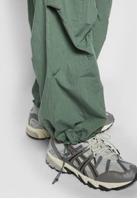 Pantalons verts amples avec des poignets élastiques, portés sur des chaussures de sport grises avec des accents gris et noirs et des lacets légers.