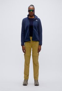 Μπλε φλις μπουφάν, μαύρο τοπ, κίτρινο παντελόνι με αποσπώμενα μέρη και γκρι παπούτσια πεζοπορίας. Μεγάλα αντανακλαστικά γυαλιά ηλίου ολοκληρώνουν την υπαίθρια στολή.