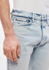 Svijetloplave traperice uskog kroja s etikietom Calvin Klein na stražnjem džepu i suptilnim prugastim teksturama na tkanini.