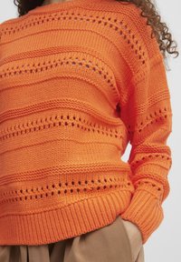 Pull en tricot orange avec des poignets et un ourlet côtelés, présentant des rayures horizontales et des détails ajourés. Texture douce et coupe décontractée.