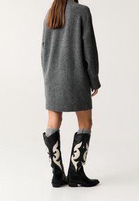 Robe-pull grise en maille, coupe oversized. Bottes noires montantes jusqu'au genou avec des motifs tourbillonnants blancs et un accent de chaussette grise en haut.