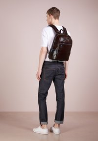 Mochila de cuero marrón oscuro con acentos azules, forma rectangular, textura suave, bolsillo frontal con cremallera y correas de hombro acolchadas.
