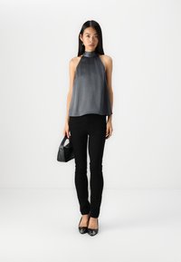 Jeune femme avec de longs cheveux portant un haut gris sans manches, un pantalon noir, des talons noirs, et tenant un sac à main noir sur fond blanc.