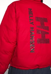 Rode pufferjack met zwart "HELly HANSEN" logo en streeppatroon op de achterkant. Heeft een gestructureerde afwerking en een verstelbare koordonderkant.