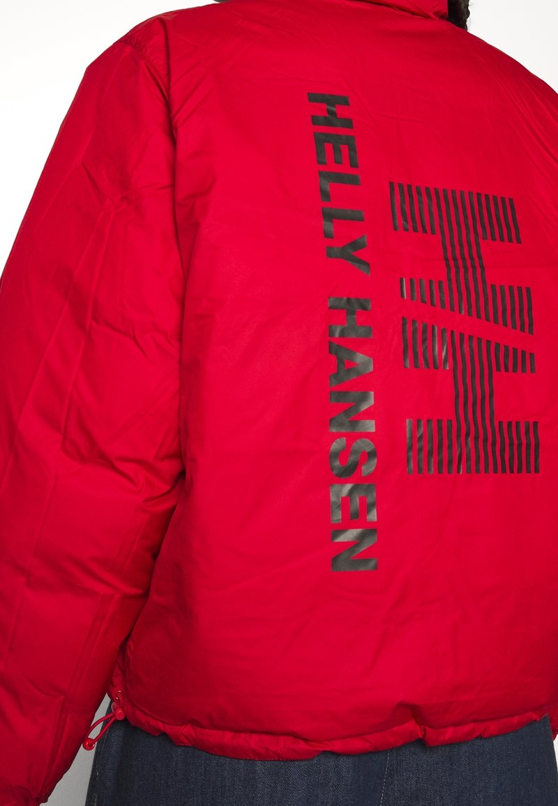 Rode pufferjack met zwart "HELly HANSEN" logo en streeppatroon op de achterkant. Heeft een gestructureerde afwerking en een verstelbare koordonderkant.