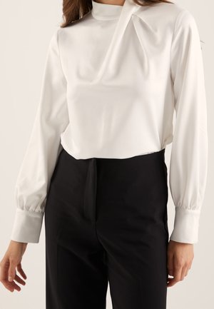 Femme portant un chemisier blanc à manches longues avec un col montant et un subtil détail de plis, associé à un pantalon noir taille haute.