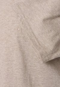 Chemise courte beige clair en tissu texturé. Présente une couture brodée et un petit logo sur la manche.