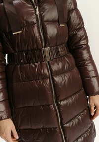 Hnědý péřový kabát s lesklou texturou, přední zip, stažený v pase s metalickým akcentem a ramenními pásy. Sekce jsou prošívané.