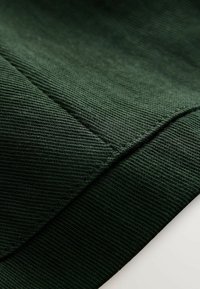 Ciemnozielona prążkowana tkanina ukazująca szczegółowe szwy i złożony brzeg, podkreślająca teksturę i konstrukcję ubrania.