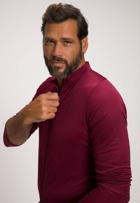 Burgundy zip-up långärmad topp i en strukturerad tyg. Har en hög krage och en liten svart logotyp på bröstet.