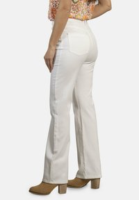 Pantalones blancos acampanados con una textura suave, cintura media y dos bolsillos traseros. Llevados con botas de tacón alto color bronceado y una blusa estampada.