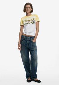 Vit t-shirt med gula gradientärmar som pryder en grafisk tryck av byggnader och palmträd. Tillsammans med avslappnande blå jeans.
