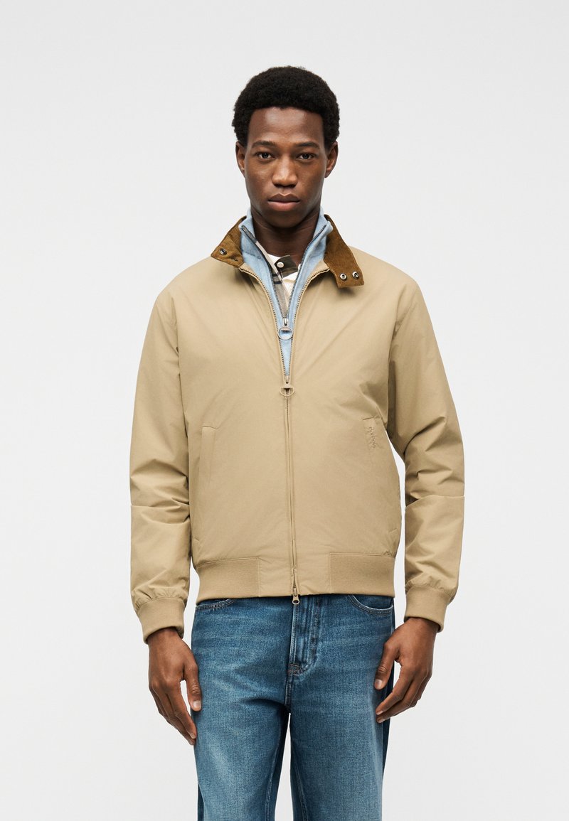 Μαύρος άντρας που φοράει μπεζ μπουφάν με φερμουάρ και καφέ γιακά, ανοιχτό μπλε εσωτερικό στρώμα και μπλε τζιν, στέκεται μπροστά σε απλό ανοιχτό φόντο.