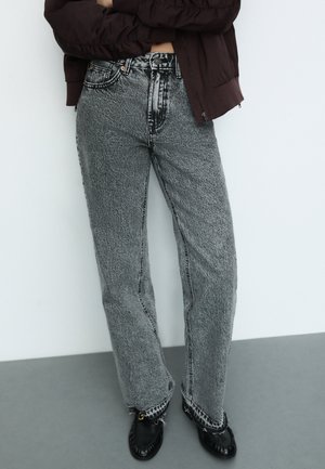 Visoke, široke traperice črne in bele teksturirane tkanine, z kontrastnim šivanjem in raztrganimi detajli na robu. V kombinaciji z črnimi čevlji.