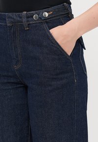 Hand resting in front pocket of dark blue denim jeans with double silver buttons and contrast stitching.