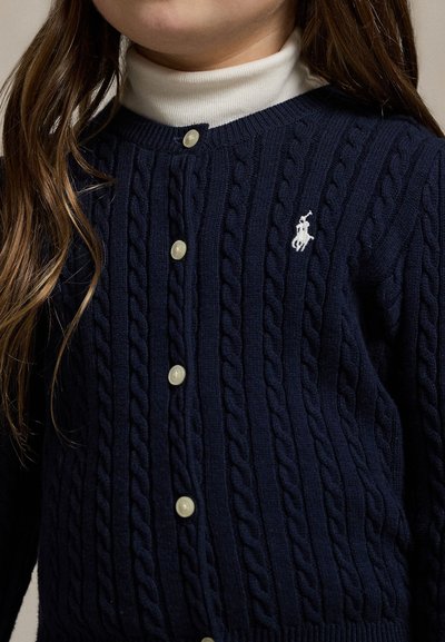 Bambino che indossa un cardigan blu navy lavorato a trecce con bottoni bianchi sopra una maglia a collo alto bianca, con un logo ricamato sul petto.