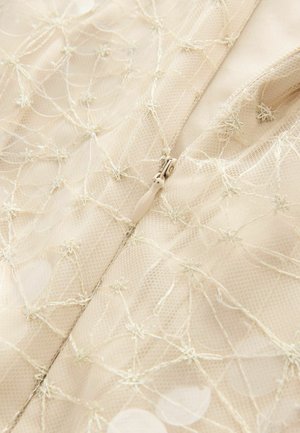 Beige nät tyg med broderade detaljer och dragkedjestängning, med cirkulära accenter och en texturerad yta.
