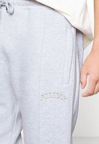 Světle šedé teplákové kalhoty z měkké látky s běžným střihem. Obsahují boční kapsy a na přední straně je vyšité "PECADOR" v tmavší šedé barvě.