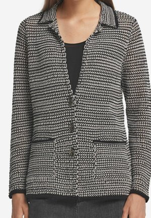 Femme portant un cardigan en maille texturée noir et blanc à boutons, avec col et deux poches avant, par-dessus un haut noir.