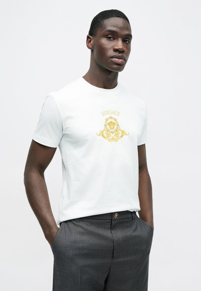Biały bawełniany t-shirt z złotym logo "Versace" i misternym wzorem lwa z przodu, w zestawieniu z szarymi spodniami.