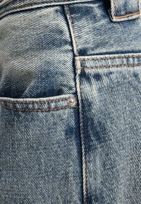 Detalle de tejido de mezclilla que muestra un bolsillo con un botón plateado. La textura presenta un degradado de azul claro a azul oscuro y costuras visibles.