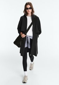 Femme portant un manteau noir, une chemise blanche, un sweat rayé noué à la taille, des leggings noirs, des baskets, des lunettes de soleil, et portant un sac à épaule noir en marchant.