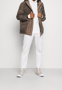 Brązowa pikowana kurtka z kapturem i wieloma kieszeniami, w połączeniu z białą bluzą z kapturem i białymi spodniami. Na nogach beżowe wysokie trampki.