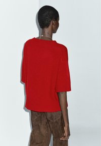 Maglione rosso lavorato a maglia con maniche corte, texture a costine e vestibilità rilassata, abbinato a pantaloni in suede marroni. Nessun accessorio notevole visibile.