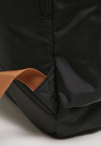 Sac en tissu noir avec une surface texturée, doté d'une sangle tissée marron attachée sur le côté et d'une ouverture zippée sur le dessus.