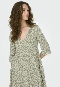 Varrukateta, V-kaelusega kleit tuhmrohelises värvitoonis, millel on tekstuuriga ikaatmuster. Kleit sisaldab kolmveerandi pikkuseid varrukaid ja pitsaktsente varruka servades.