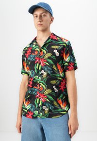 Jonge man met een blauwe pet, een zwart shirt met korte mouwen en een kleurrijk tropisch bloemenpatroon, en lichtblauwe spijkerbroek tegen een witte achtergrond.