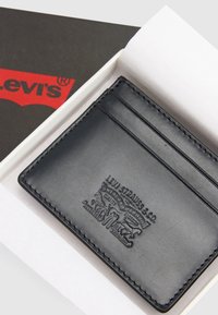 Μαύρος δερμάτινος θήκη καρτών Levi's με ανάγλυφο λογότυπο, διαθέτει τρεις θήκες για κάρτες, τοποθετημένη μέσα σε λευκό κουτί με μερικώς ορατή κόκκινη και μαύρη ετικέτα Levi's.