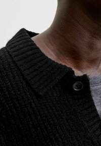 Juodas megztinis cardigan su raukšlėtu apykakle, turintis mygtuko užsegimą. Tekstūra atrodo minkšta ir pūkuota, uždėta ant pilkos marškinėlių.