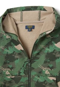Giacca verde mimetica con zip e foderatura in pile beige. Presenta una chiusura con cerniera e un'etichetta interna che recita "OVS kids". Tessuto morbido al tatto.