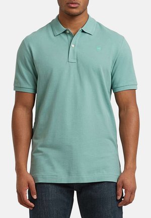 Homme portant un polo vert clair à manches courtes avec deux boutons, associé à un jean bleu foncé, debout devant un fond blanc.