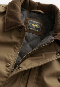 Veste marron avec col en velours côtelé, doublure matelassée grise, fermeture éclair en métal, boutons-pression, et étiquette noire portant l'inscription "1982 NEXT Durable Goods".