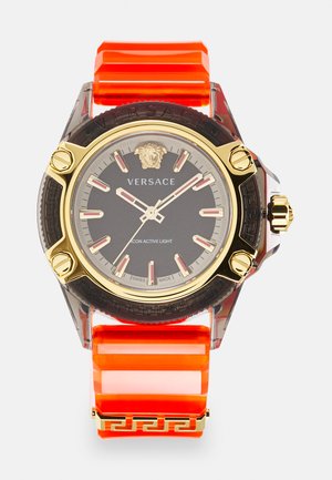 Versace watch with a black dial, gold accents, and a transparent case. Features a bold orange rubber strap and Greek key design details.