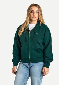 Sweat à capuche vert zippé avec un cordon de serrage, poignets côtelés et un petit logo sur la poitrine. Associé à un jean bleu clair. Matériau doux.