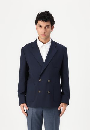 Navy blue double-breasted blazer with two front pockets, bronze buttons, and a structured design. Worn over a white shirt.