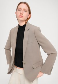 Beige skreddersydd blazer med smal passform, spiss krage, enkel knappelukking og frontlomme. Brukt over en svart polo-genser. Glatt tekstur.
