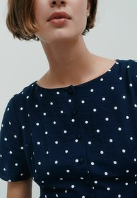 Marinblå blus med vita prickar, rund halsringning, korta ärmar och knapprad framtill. Mjuk textur.