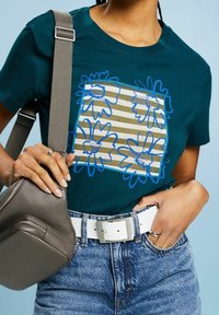 Teal bomullströja med blå blommig grafik ovanpå randigt mönster. En vit bälte och en grå axelväska kompletterar looken.