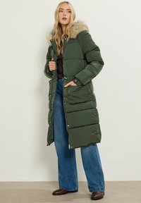 Abrigo acolchado verde con capucha de piel sintética, diseño acolchado y bolsillos laterales. Combinado con jeans de pierna ancha y botas marrones.