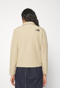 Beige jacka med mjuk textur, utrustad med krage och långa ärmar. Bakom finns en svart logotyp från The North Face.
